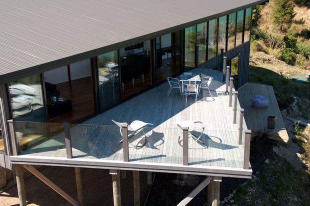 Elevated view of a modern, single-storey building integrated into a steep natural hillside. A broad timber deck with glass and dark-framed balustrades extends along full-height glazing, with outdoor dining and lounge furniture. The building has a low-pitched roof, dark structural framing, and extensive glass overlooking the rugged site.