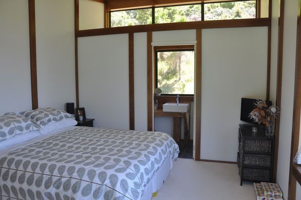 A bright bedroom with a double bed dressed in a grey and white leaf-patterned duvet, dark timber framing, pale wall panels, and high clerestory windows. A compact open vanity area with a white basin is visible through the doorway, along with a dark chest of drawers and decorative arrangement. The room overlooks leafy greenery outside.