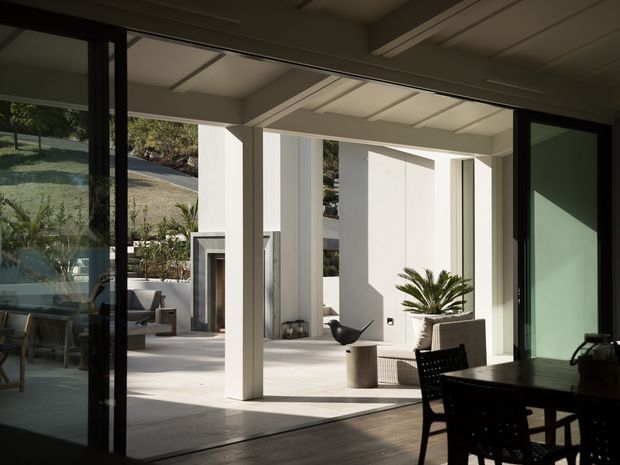 View from a dark, glazed interior dining area through wide black-framed sliding doors to a bright covered outdoor living terrace. The terrace features a white structural colonnade, exposed-beam ceiling, pale hardscape flooring, a built-in fireplace, lounge seating, a sculptural potted palm, and a landscaped sloping site beyond. The scene has strong indoor-outdoor connection and crisp modern detailing.