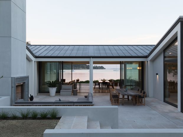 A contemporary courtyard-facing outdoor living area of a low-profile home, with expansive floor-to-ceiling glazing opening toward a calm waterfront view. Pale rendered walls, a dark standing-seam metal roof, a built-in fireplace, paved terraces, timber outdoor dining furniture, and a sunken central courtyard create a restrained indoor-outdoor living arrangement.
