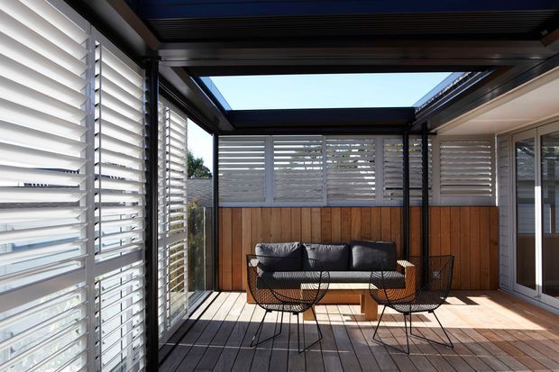 A stylish covered outdoor terrace or sunroom with timber decking, black-framed glazing, timber wall panelling, and extensive white horizontal louvre screens. The space contains a compact outdoor lounge setting with a sofa, coffee table, and wire-frame chairs. A partially open overhead structure brings in strong sunlight and creates a bright indoor-outdoor atmosphere.