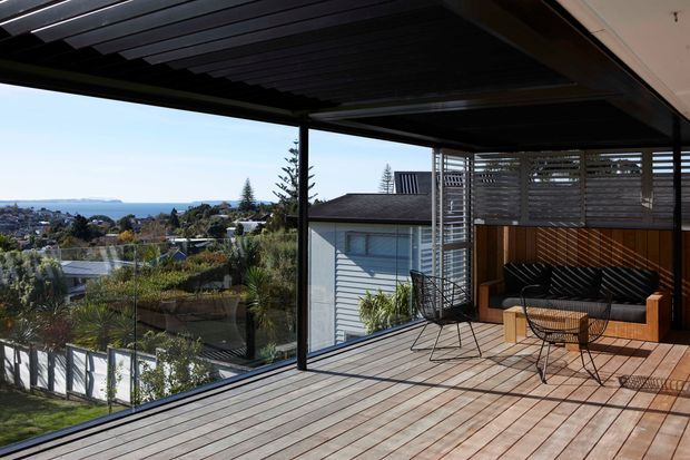 A contemporary covered outdoor terrace overlooking a suburban coastal landscape. The spacious deck features timber flooring, dark metal framing and ceiling slats, frameless glass balustrades, a built-in timber bench with black cushions, and a small outdoor dining setting. A slatted timber privacy screen encloses part of the terrace, with the ocean visible in the distance.