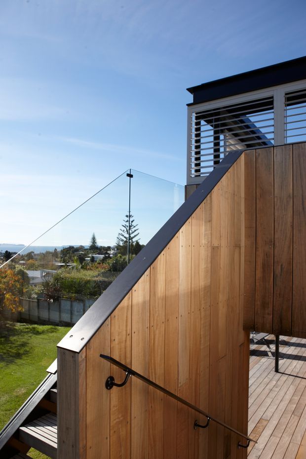 Exterior view of a contemporary multi-level residential deck and stairway. Warm vertical timber screening and decking frame an open stair with a dark metal handrail, while clear frameless glass balustrades provide expansive suburban and coastal views. The upper level features white-framed horizontal louvres beneath a dark flat roof edge, with bright blue sky and landscaped surroundings beyond.