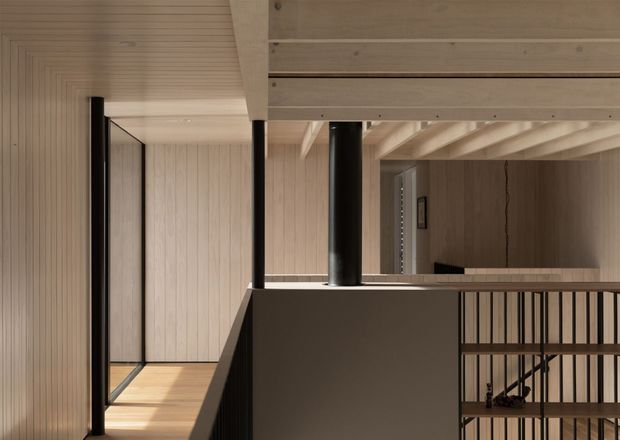 Interior view of a refined modern home focused on a stair landing and upper-level circulation space. Pale timber-lined walls and ceiling, exposed light timber beams, black cylindrical columns, and a dark balustrade create a warm yet highly restrained composition. A stair descends behind vertical black metal railing, while a glazed opening and timber floor extend along the left side.