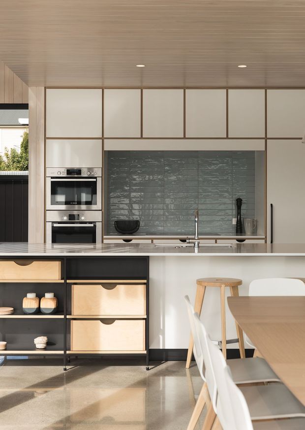 A contemporary open-plan kitchen and dining area with pale timber-lined ceiling and cabinetry, dark open shelving with light timber drawers, integrated stainless-steel ovens, a grey textured tile splashback, and a pale stone island benchtop. A timber dining table and white chairs occupy the foreground, with warm natural light entering from the side.