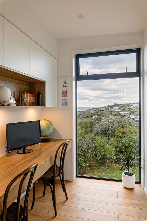 Compact, light-filled home office with a built-in timber desk, computer monitor, dining-style chairs, and integrated white overhead cabinetry with an open timber display niche. A full-height dark-framed glazed door or window provides expansive views over a leafy residential hillside and water beyond. The room has pale timber flooring, white walls and cabinetry, and warm wood accents.