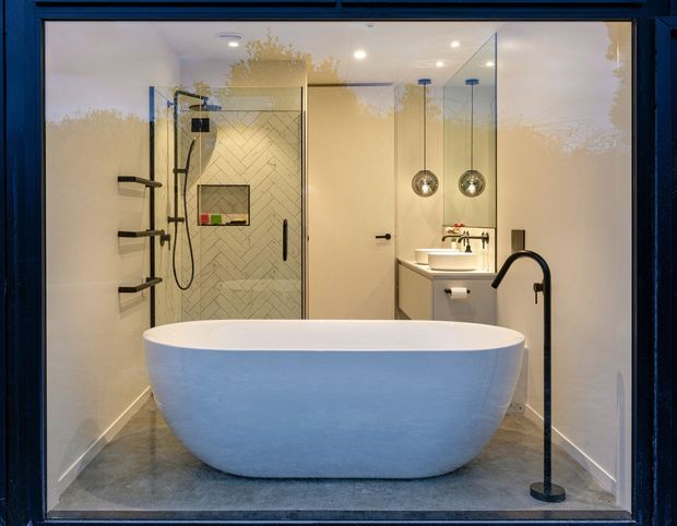 A refined contemporary bathroom viewed through dark-framed glazing, featuring a freestanding white bathtub in the foreground, a frameless glass walk-in shower with herringbone wall tile, floating vanity with vessel basin, pendant lights, and matte black tapware and fixtures. The room has warm neutral walls, grey flooring, and a clean, softly lit atmosphere.