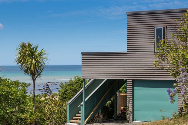 Exterior view of a contemporary multi-level coastal house overlooking the sea. The building has horizontally lined dark timber-toned cladding, a raised form with an open stairway beneath, turquoise painted lower walls, and simple rectangular windows. Coastal vegetation and a beach are visible around the property.