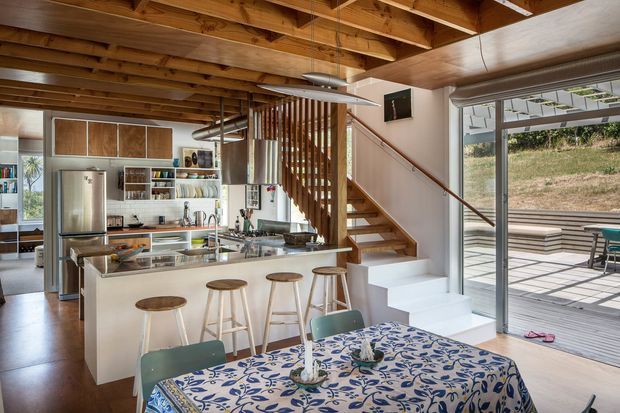 Open-plan kitchen and dining area in a warm timber-lined home. The kitchen features a white island with integrated sink and raised bar seating, stainless-steel appliances, open shelving, and timber cabinetry. A timber stair with vertical slat balustrade leads upward beside the kitchen. Exposed timber ceiling joists and timber ceiling boards add strong architectural character, while large sliding glass doors open to a paved outdoor terrace. A patterned dining table occupies the foreground.