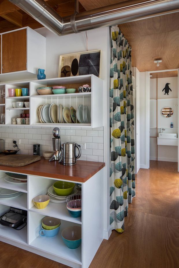 A compact, colourful kitchen with open white shelving displaying crockery and bowls, a warm timber benchtop, white subway-tile splashback, and timber flooring. Exposed stainless-steel ductwork runs beneath a timber-lined ceiling, giving the space an informal industrial character. A patterned curtain screens an adjacent hallway or room, with a bathroom visible beyond.