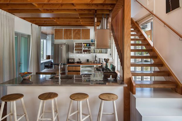 A bright, compact open-plan kitchen viewed from the breakfast bar, with four timber stools, a dark stone island benchtop, stainless-steel sink and appliances, white cabinetry and tiled splashback. Exposed timber ceiling beams and a timber open-riser staircase with vertical slatted screening define the space, while glazing brings in natural light.