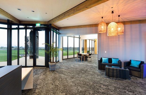 A spacious contemporary commercial reception or lounge area with extensive black-framed floor-to-ceiling glazing, exposed timber beams, patterned carpet, and a seating arrangement beneath three woven pendant lights. A smaller dining or meeting area is visible beyond the open glazed partition, with expansive green grounds outside.