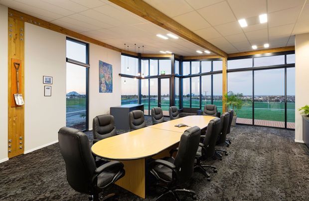 A bright contemporary boardroom with a large oval timber meeting table surrounded by black leather office chairs. Floor-to-ceiling glazing provides expansive views toward a sports field and distant landscape. The room features exposed timber beams, suspended pendant lights, acoustic carpet tiles, white ceiling tiles, and a glazed corner facade.