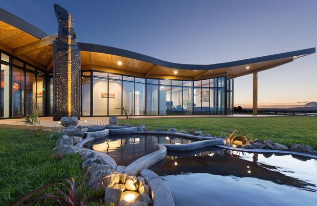 A low, expansive contemporary building with a dramatic curved roofline and extensive floor-to-ceiling glazing, viewed at dusk from a landscaped exterior. A tall carved sculptural column marks the entrance, while a naturalistic rock-edged reflecting pond and lawn occupy the foreground. Warm recessed lighting glows beneath the timber-toned soffit, and interior furnishings are visible through the glass.