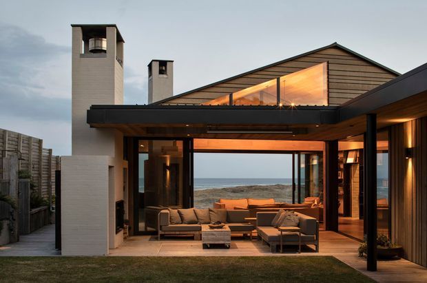 A contemporary coastal house viewed from the rear garden, featuring a sheltered outdoor living terrace opening into a warm, glass-lined living space. The building combines dark horizontal timber cladding, black-framed glazing, concrete-rendered chimney towers, and a pitched gable roof. Through the expansive glazing, a neutral-toned seating area and ocean horizon are visible, while warm interior lighting creates a welcoming evening atmosphere.