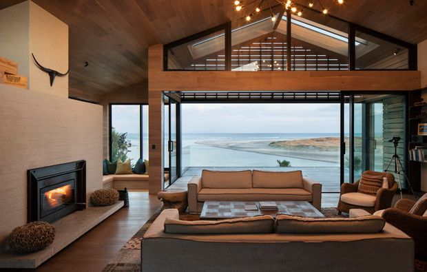 A warm, contemporary coastal living room with a dramatic double-height glazed wall overlooking the ocean and headland. The room features a large sectional sofa, additional lounge seating, a central coffee table, a built-in fireplace set into a pale masonry wall, dark structural framing, extensive timber ceiling and upper-level cladding, and a balcony or mezzanine with horizontal balustrading. Decorative pendant lighting and shelving add character while preserving the expansive water view.