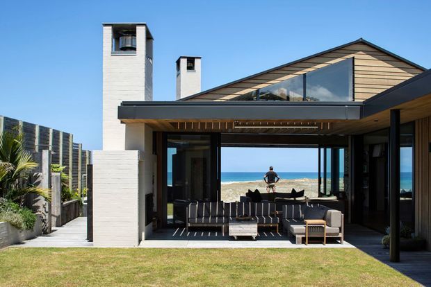 Contemporary coastal house viewed from the rear, with a sheltered outdoor living area opening directly toward the beach and ocean. The building features a strong gabled roof form, pale horizontal timber cladding, white masonry chimney volumes, dark-framed expansive glazing, and a timber-lined soffit. A lounge setting occupies the covered terrace, with lawn and landscaped boundaries in the foreground.
