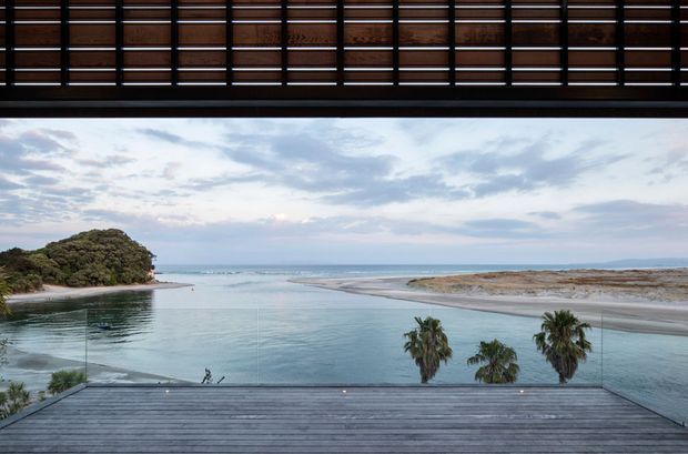 A wide architectural view from a covered outdoor terrace or balcony overlooking a calm coastal inlet, sandy shoreline, and ocean beyond. The foreground features dark horizontal timber decking and a frameless glass balustrade, while a deep overhead timber slatted screen creates a strong architectural frame. Palm trees and low coastal vegetation are visible near the water.