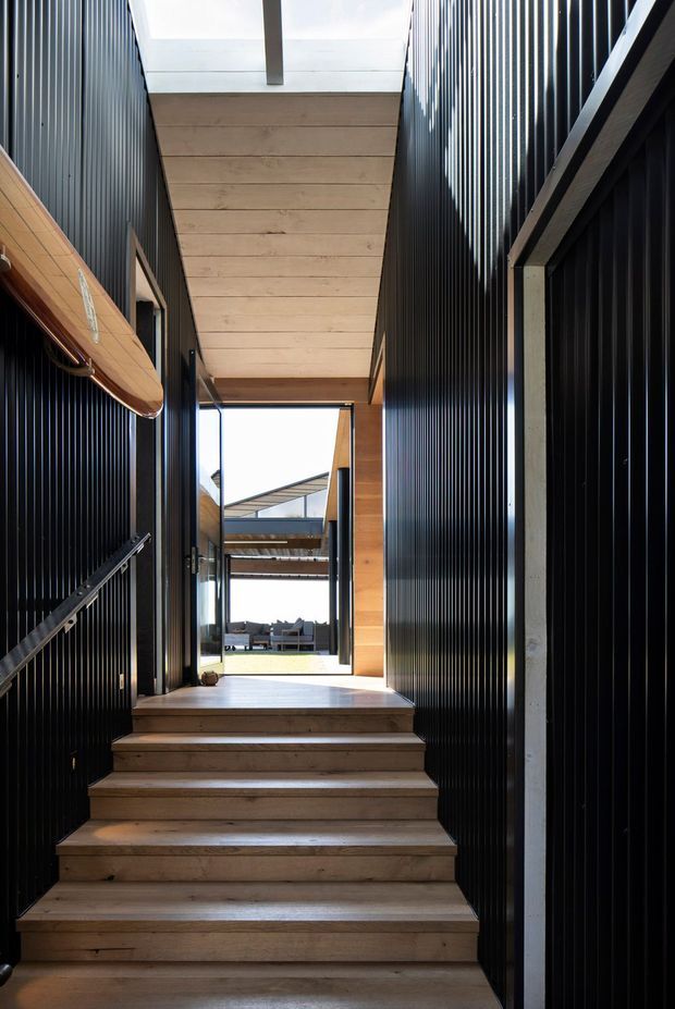 A narrow, modern interior stairway with broad timber treads rising between dark vertically ribbed wall panels. A pale timber-clad ceiling with a central skylight brings daylight into the stair hall, while a full-height glazed opening at the landing reveals an outdoor area and adjacent roof forms. A timber surfboard is mounted along the left wall as a decorative feature.