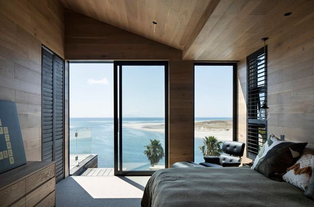 A warm, wood-lined coastal bedroom with a low pitched ceiling and expansive floor-to-ceiling glazing overlooking the ocean and sandy shoreline. The room contains a large bed with dark bedding, a timber drawer unit, a black lounge chair, and dark louvred window screens. Natural timber surfaces dominate the ceiling and walls, creating a calm, contemporary retreat with abundant daylight and strong indoor-outdoor connection.