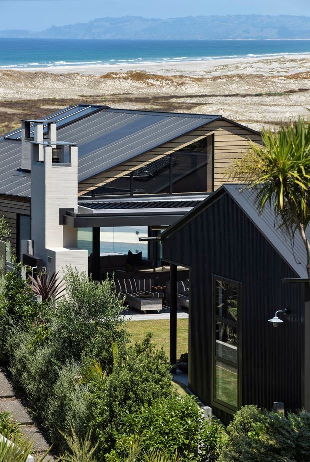 Exterior view of a contemporary coastal house overlooking a sandy beach and ocean. The home has dark standing-seam metal roofs, dark vertical cladding, pale horizontal weatherboards, large glazed openings, and a covered outdoor living terrace with built-in seating. Native and coastal landscaping surrounds the buildings.