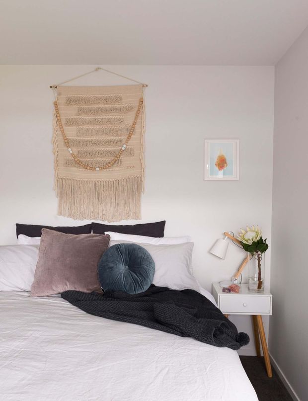 A softly styled bedroom with a white bed and layered white bedding, dark charcoal throw, mauve and deep blue cushions, and a cream macramé wall hanging above the bed. A small white bedside table with timber legs, reading lamp, vase of flowers, and framed artwork sits against pale grey walls. The room has a calm, warm, bohemian-inspired feel.