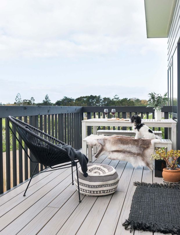 A styled elevated outdoor timber deck or balcony with dark vertical balustrades, a black woven lounge chair, a pale timber bench and table, patterned floor cushion, layered sheepskin throws, outdoor rug, and potted plants. A small dog sits on the bench, with leafy treetops visible beyond the railing. The setting is bright, relaxed, and nature-oriented.