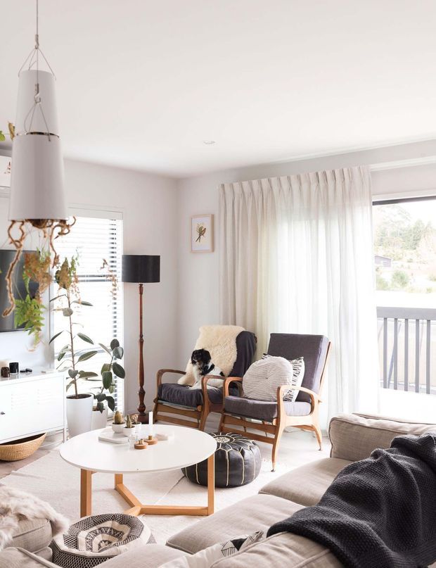Bright, airy living room with a Scandinavian-inspired arrangement of a pale sectional sofa, round white coffee table, wood-framed lounge chairs, floor lamp, indoor plants, soft textiles, and sheer curtains. Large glazing opens to a balcony, while white walls and light flooring create a calm, inviting atmosphere.