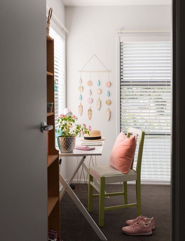 A bright, compact home office or study viewed through a partially open door. The room has white walls, dark carpet, a small folding desk with a potted plant, books and stationery, a green-painted chair with a coral cushion, white plantation-style blinds, open wooden shelving, and playful pastel hanging wall decor. Natural daylight creates an airy, cheerful atmosphere.