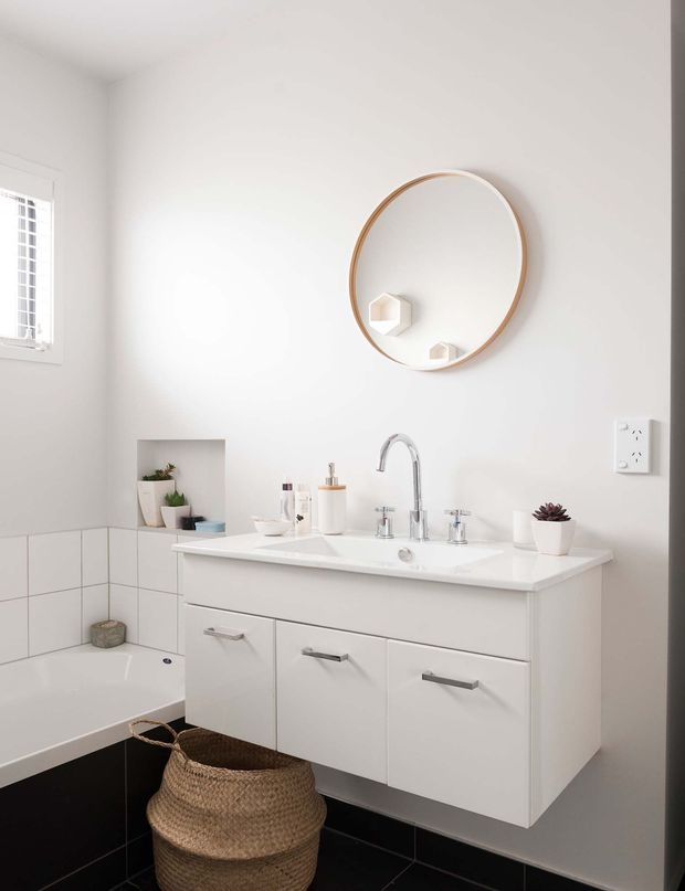 Bright, compact contemporary bathroom with a floating white vanity, integrated white basin, chrome tapware, round timber-framed mirror, white wall tiles, and black floor and lower wall tiles. A bathtub, small plants, toiletries, and a woven basket complete the clean, minimalist fit-out.