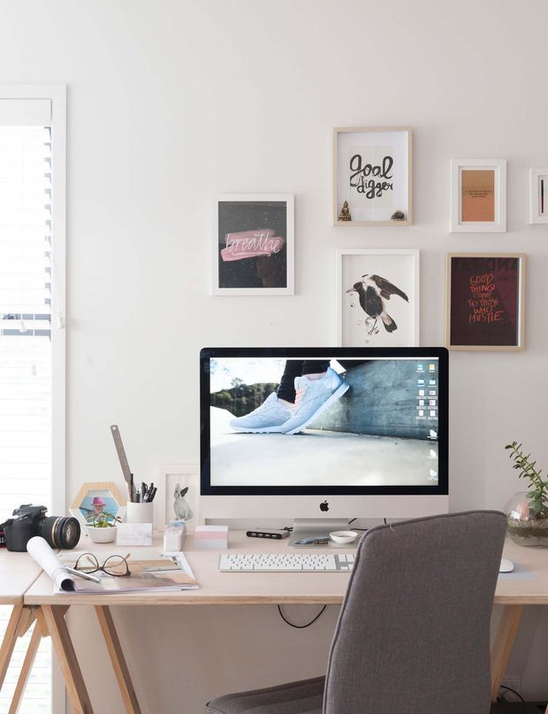 A bright, compact home office with a pale timber desk, desktop computer, keyboard, stationery, camera, papers and an upholstered desk chair. White walls feature a gallery arrangement of framed inspirational artwork, while a window with white blinds provides natural light.