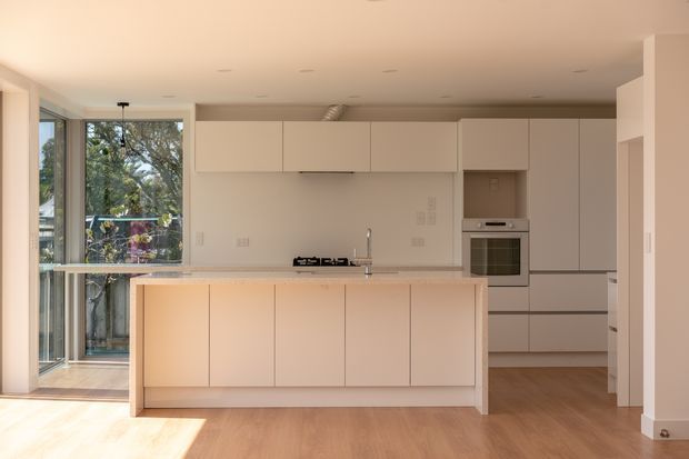 A bright, minimalist contemporary kitchen viewed straight on. The room features a large pale island with an integrated cooktop and sink, extensive handleless light cabinetry, a built-in oven, pale stone-look benchtops, and light timber flooring. Floor-to-ceiling glazing at left opens toward a leafy exterior and brings in strong natural light.