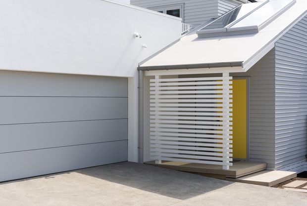 Exterior view of a compact contemporary residential entry and attached garage. The facade uses pale grey horizontal weatherboard cladding, a wide sectional garage door, and a covered entry porch screened by horizontal white slats. A bright yellow-green front door adds a strong accent, while a small rooflight/skylight is visible above the entry canopy. Concrete paving forms the driveway and entry approach.