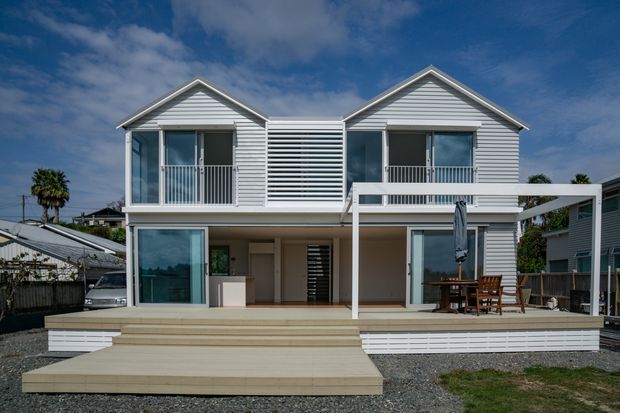 Front-facing exterior photograph of a contemporary coastal-style two-storey home with two prominent gabled wings, white horizontal weatherboard cladding, balconies, extensive glazing, and a broad timber deck with steps. A central horizontal louvred screen links the upper volumes, while a covered outdoor dining area occupies the right side. The house sits in a low-density suburban setting under a partly cloudy sky.