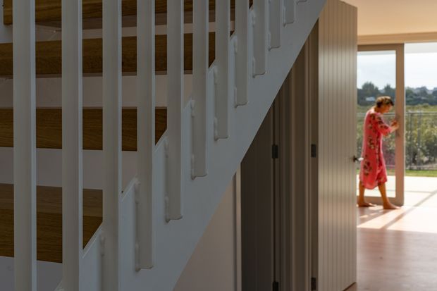 Interior view focused on a contemporary staircase with white vertical balustrade slats and exposed warm timber stair treads. A tall ribbed timber-toned built-in partition or cabinetry runs beneath and beside the stairs, leading toward a bright glazed living area. A person in a red dress is visible in the background, which opens to an outdoor landscape.