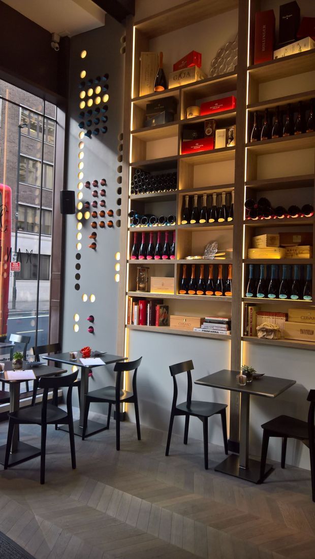A refined wine bar or café interior with tall illuminated timber display shelving filled with wine bottles, books, boxes and glassware. Small black café tables and chairs are arranged beside a large industrial-style framed window, while decorative circular bottle displays create a feature wall. The space has warm accent lighting, dark walls and a herringbone timber floor.