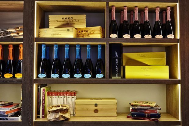 Close-up of a dark timber built-in shelving unit with warm integrated lighting. The shelves display champagne bottles, branded boxes, books, and a small wooden storage box, creating a refined wine-storage and display feature.