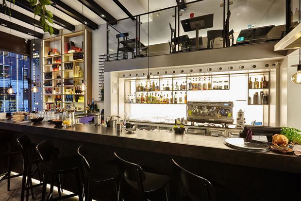 A sophisticated commercial bar and dining venue with a long dark countertop, black bar stools, illuminated back-bar shelving filled with bottles and glassware, a professional espresso machine, pendant lighting, exposed black ceiling beams, and a glass-fronted mezzanine above. The interior combines dark finishes with warm timber shelving and bright integrated lighting.