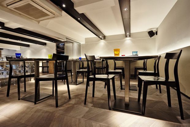 A modern restaurant or café dining interior with multiple dark timber tables, tall black chairs, and a long communal-style table arranged against a white wall. The space features exposed black ceiling beams with integrated spotlights, warm wood-look flooring, a partial slatted divider, and subdued ambient lighting.
