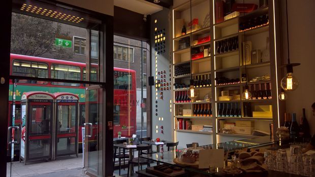 A dimly lit urban bar or restaurant interior viewed from the dining area, with floor-to-ceiling street-facing glazing, a built-in illuminated shelving wall displaying bottles and glassware, pendant lights, and small tables. A city street with red buses and a public telephone booth is visible outside.