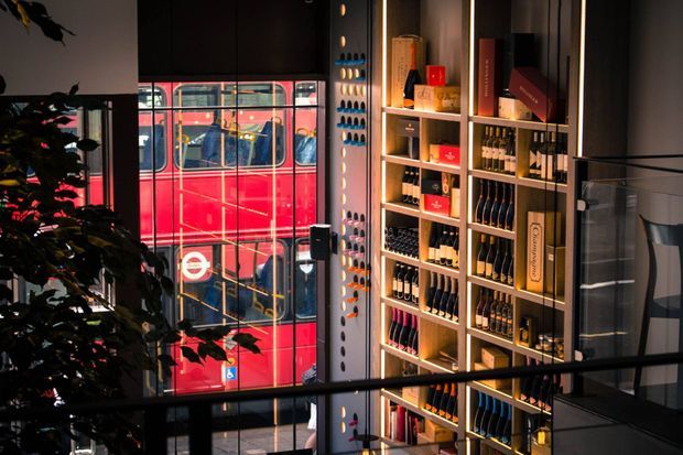 A stylish commercial retail interior with tall illuminated shelving displaying bottles and packaged products, dark wall finishes, and a large window overlooking a red double-decker bus. A leafy indoor plant and black metal balustrade add visual depth.
