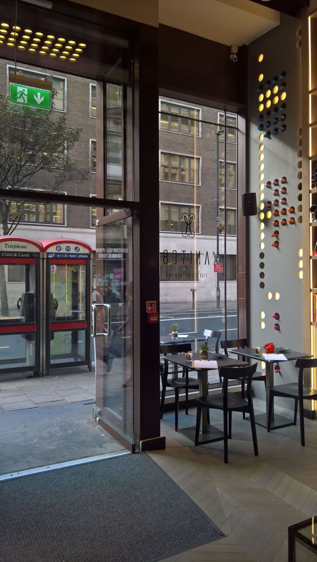 Interior view of a contemporary café or restaurant entrance, with full-height glazed frontage, dark timber or painted framing, small dining tables and chairs, and a distinctive perforated feature wall illuminated by warm circular lights. The herringbone-patterned light floor and dramatic dark ceiling create a refined urban hospitality setting. A street-facing telephone kiosk and neighbouring brick building are visible through the glazing.