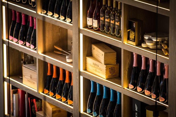 A close-up view of a dark timber built-in wine display and storage wall, with illuminated open shelving holding numerous wine bottles, wooden wine crates, glasses, and small wine-related accessories. The warm integrated lighting and rich wood tones create an intimate, upscale wine-storage atmosphere.