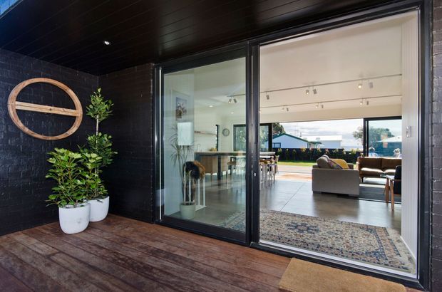 A modern indoor-outdoor living connection viewed from a covered timber deck. Black-painted brick walls and ceiling frame large sliding glass doors opening into a bright open-plan living and dining area, with a kitchen visible beyond. Potted greenery and a circular timber wall feature add warmth, while a patterned rug and neutral upholstered seating create a relaxed contemporary setting.