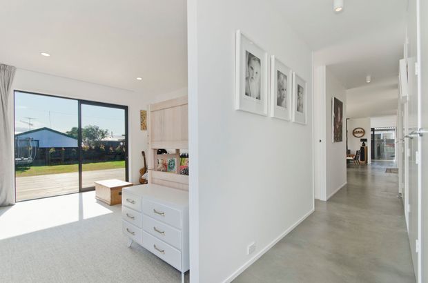Bright contemporary interior showing a hallway opening onto a carpeted bedroom or living space, with a large sliding glass door to a timber deck, built-in white cabinetry, framed artwork, and polished concrete flooring extending through the corridor.