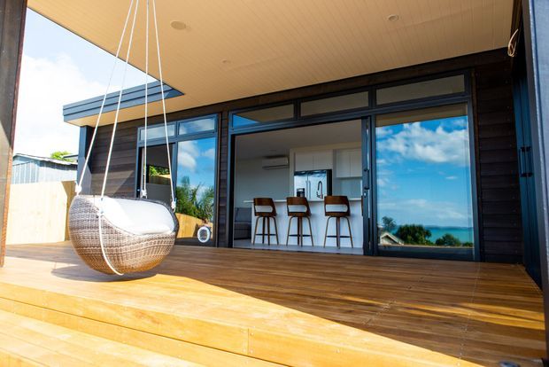 Bright exterior view of a contemporary coastal home, showing a timber deck and broad covered outdoor living area with a hanging egg chair. Large dark-framed glazed doors open into a kitchen with a small island or breakfast bar and three stools. The ocean and sky are visible beyond the property.