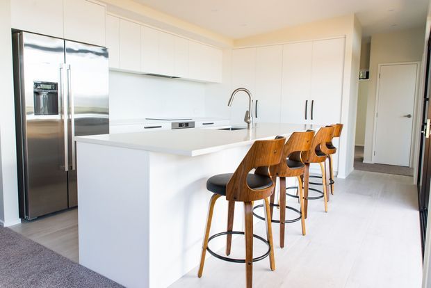 Bright contemporary kitchen with extensive white flat-panel cabinetry, a large white island with integrated sink and gooseneck tap, stainless-steel French-door refrigerator, and four curved wooden bar stools with black seats. Pale flooring and neutral walls create a clean, airy interior.