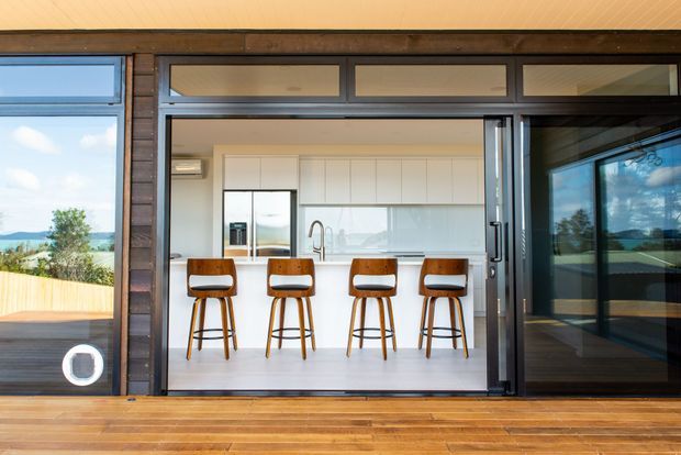View from a timber deck through open glazed sliding doors into a bright contemporary kitchen. The kitchen features a long white island with integrated sink and four wooden bar stools, white full-height cabinetry, stainless-steel appliances, and large windows overlooking a coastal or rural landscape. Dark timber-framed glazing and warm timber decking surround the opening.
