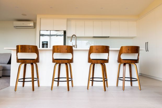 Bright contemporary kitchen with a long white island and four sculptural timber bar stools. White full-height cabinetry, integrated appliances, a stainless steel gooseneck tap, pale timber flooring, and a partial living area create a clean, open-plan interior.