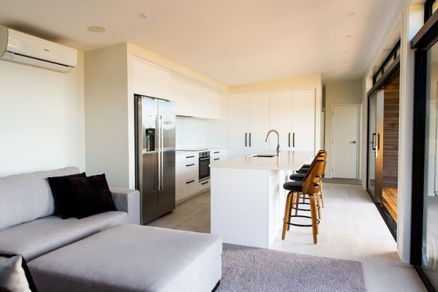 Bright open-plan kitchen and living area with white full-height cabinetry, a central island with integrated sink, stainless-steel appliances, timber bar stools, and a light grey sectional sofa. Large black-framed glazed doors line one side of the room, creating an airy contemporary interior.